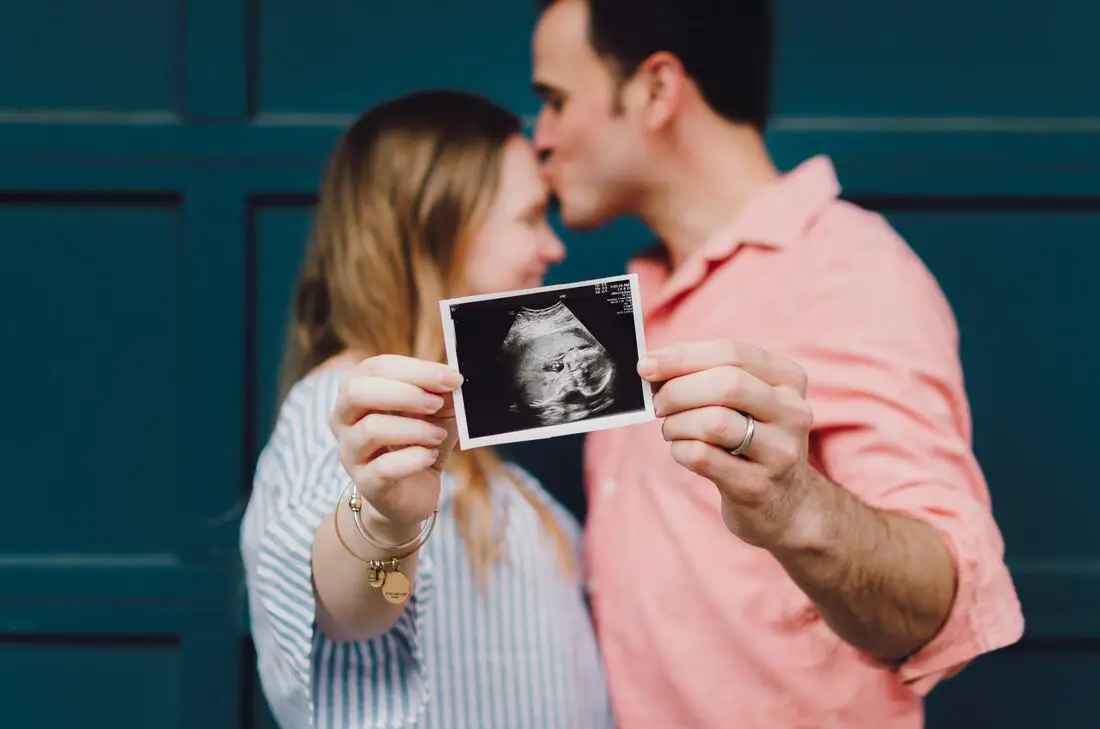 foto de casal, homem beijando a testa da mulher, com foto de ultrassom de bebê. Foto para capa do post sobre frases de gravidez para status.