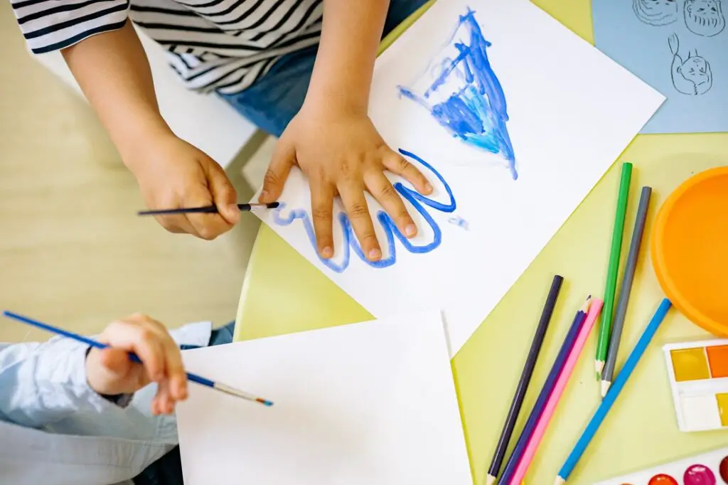 duas crianças vistas do alto, fazendo atividades de pintura, uma delas contornando a mão com uma tinta azul, atividade que estimula a coordenação motora fina.
