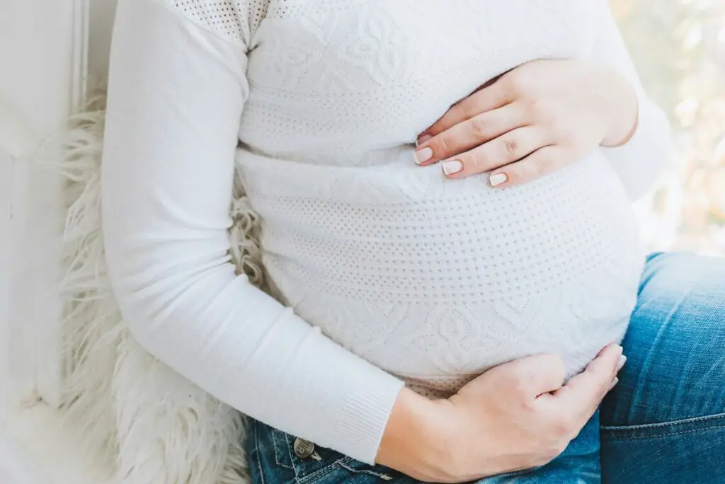 mulher gravida de blusa de camisa branca, com as mãos na barriga, dentro do post sobre calcular semanas em meses de gestação