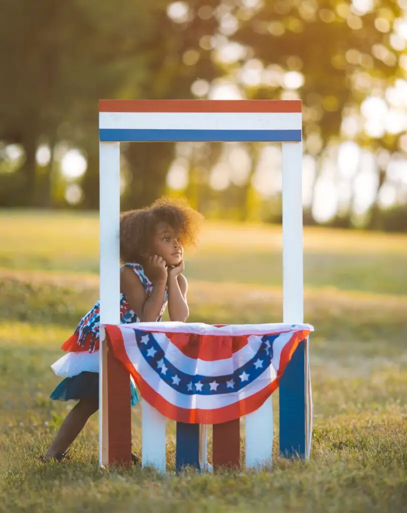 menina atras de uma estrutura de madeira, enfeitada com temas da bandeira dos Estados Unidos para ilustrar post Nomes femininos americanos.