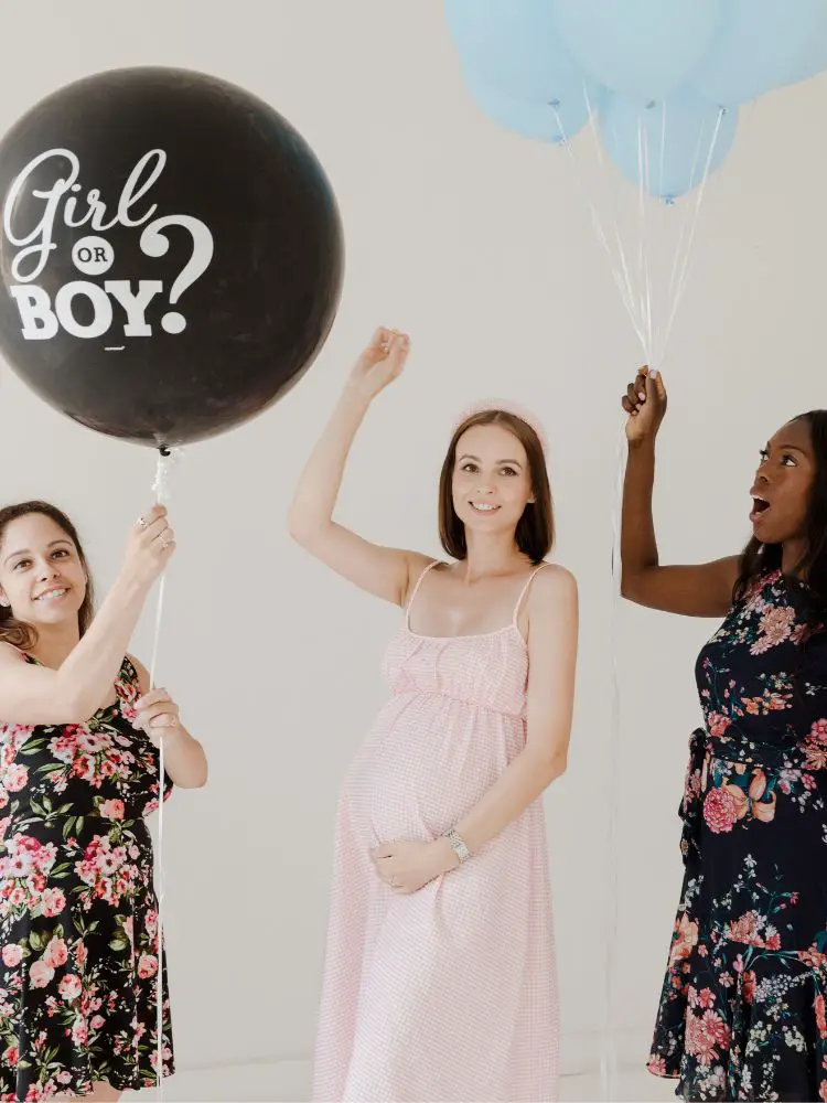 Three women at a gender reveal party, one of them holding a balloon with "boy" or "girl" written on it, the other with a spike to pop the balloon, and the third one holding blue balloons. Gender Reveal Games.