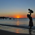 mãe segurando bebe no alto, em uma praia, no por do sol, mensagem para mãe de primeira viagem