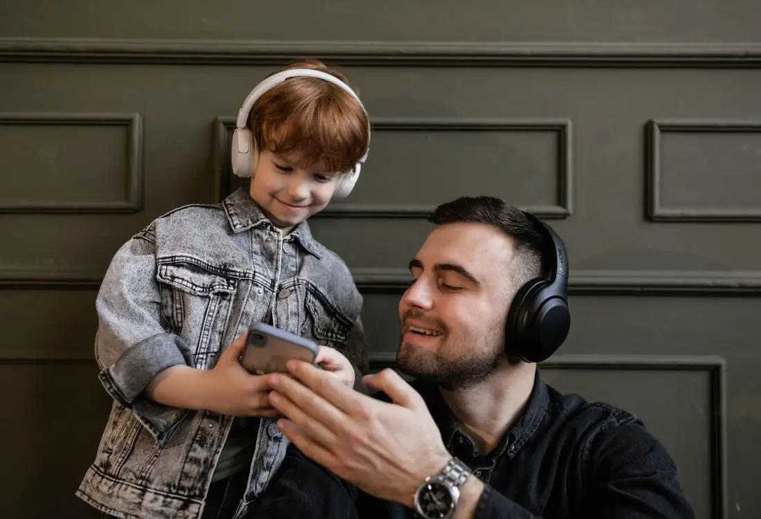 um menino ruivo e um homem mais velho de cabelo preto, ambos com fone de ouvido, ouvindo música e olhando para a tela de um celular, capa de post música para ouvir com crianças.